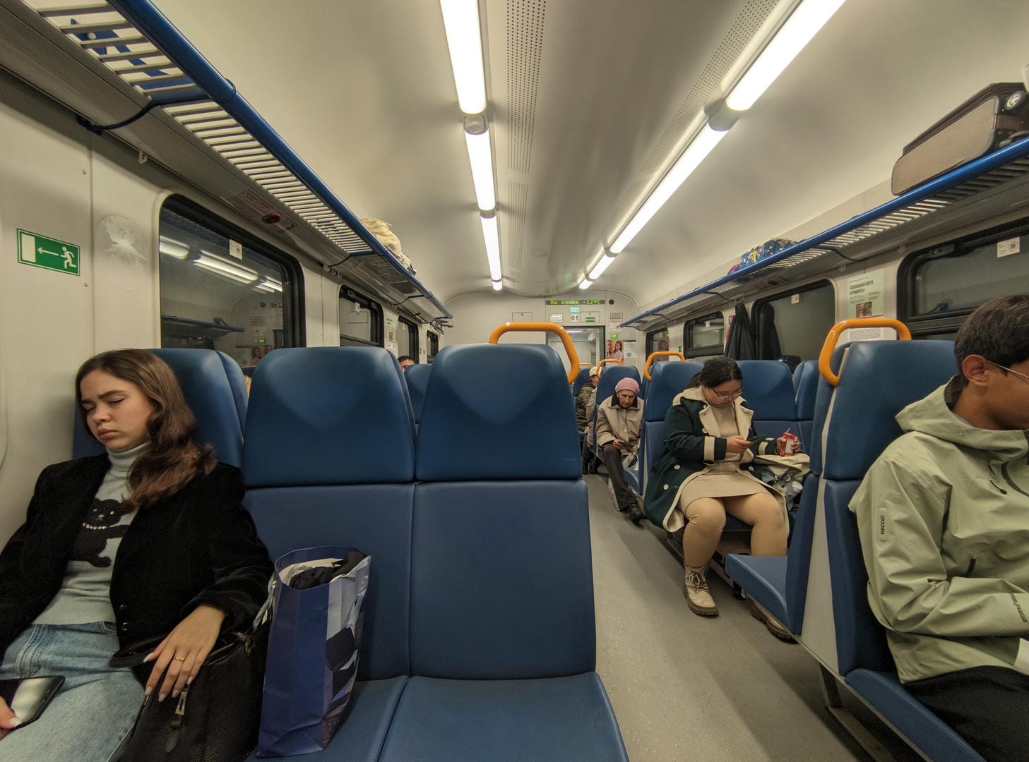 Inside a train carriage, passengers are sitting on blue seats; some are sleeping, and others are looking at their phones.