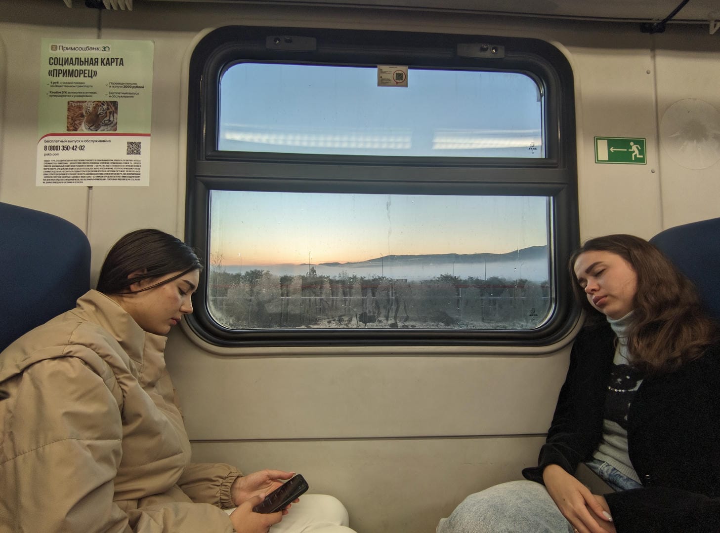 Two women sitting by the train window, the woman on the left playing with her phone, and the woman on the right leaning back in her seat to rest.
