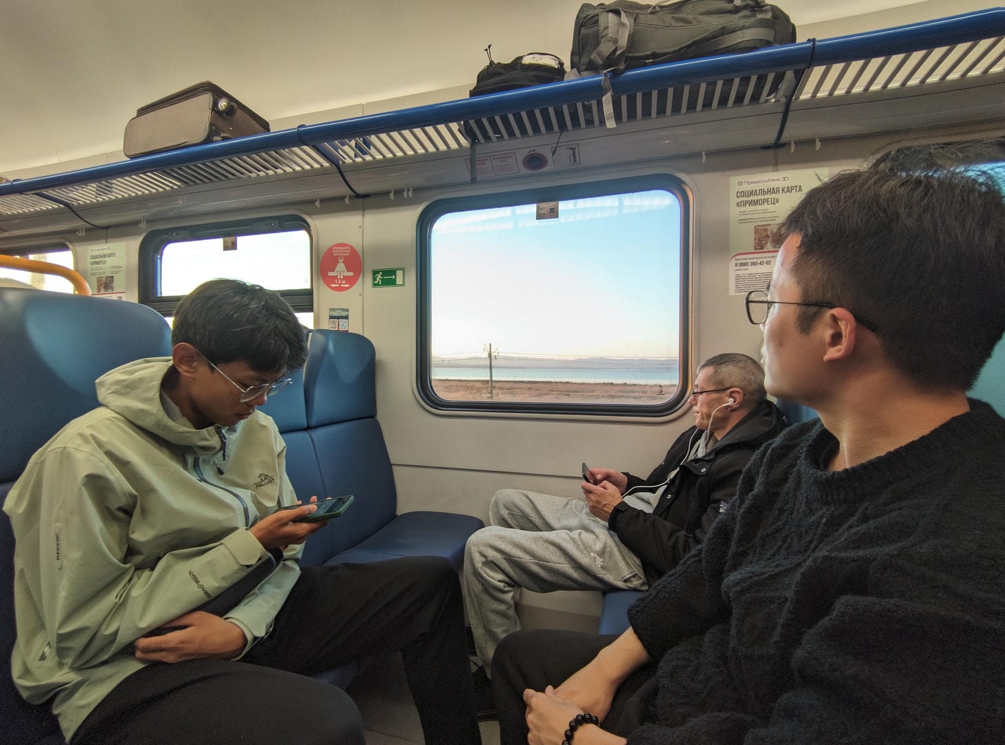 Three people sitting on a train, the person on the left wearing glasses, the person in the middle wearing headphones, and the person on the right looking out of the window.