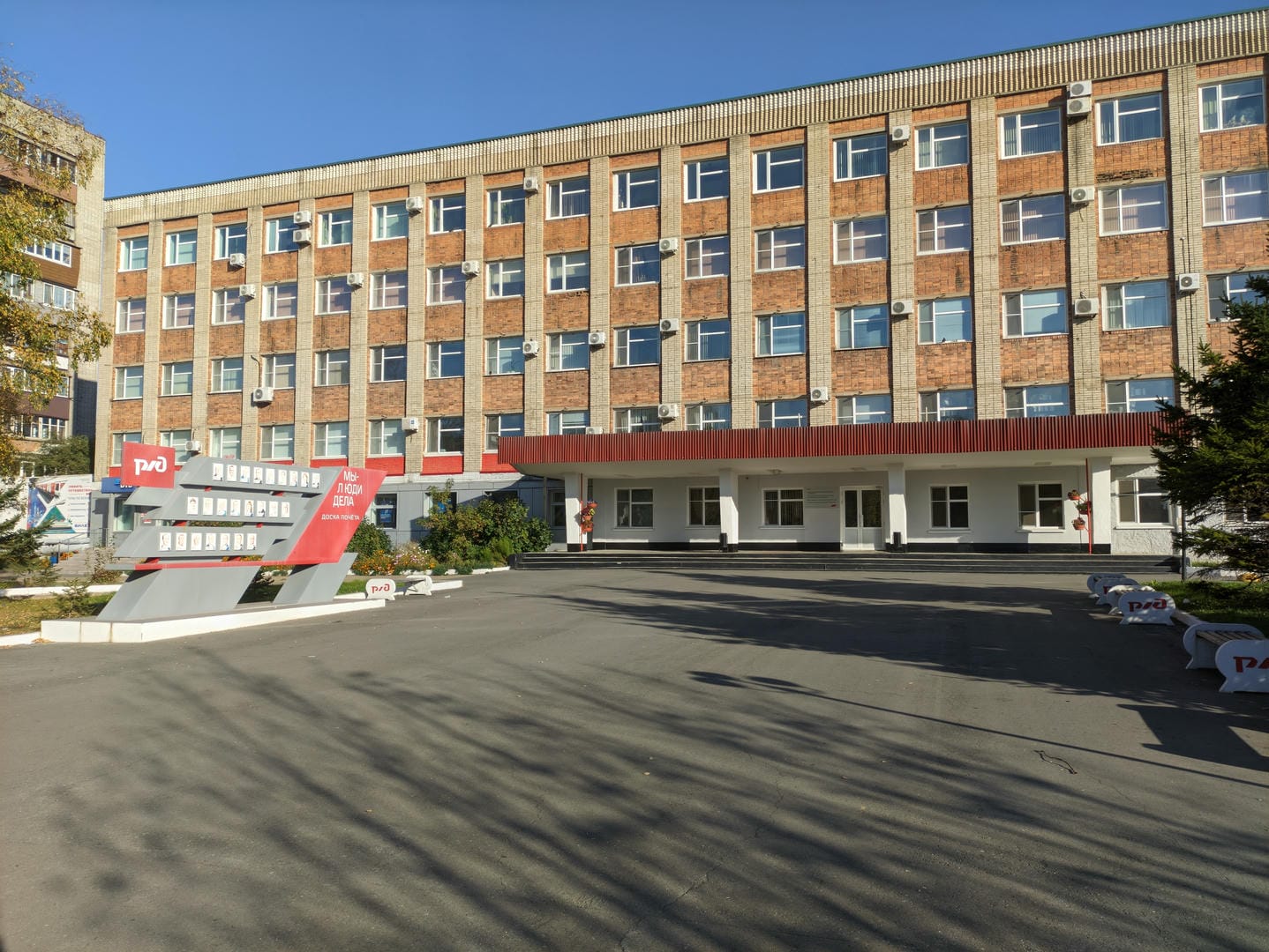 In front of a multi-story building stands a red sculpture, with air conditioning units hanging on the exterior wall.