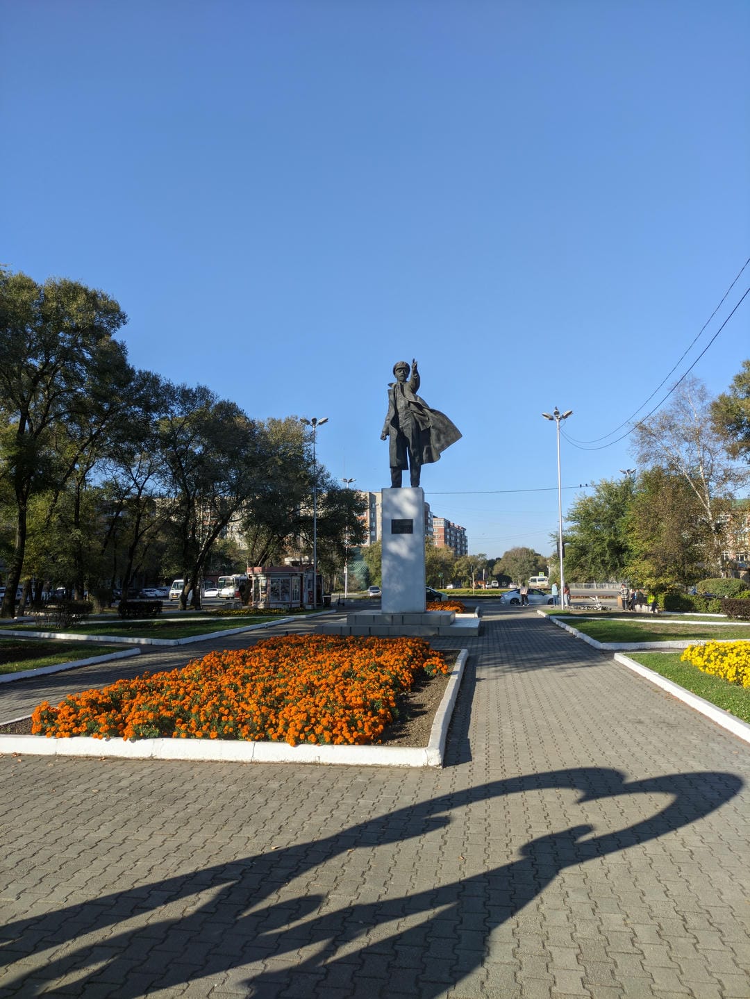 A statue stands in the center of a flower bed, with a plaque on its pedestal, surrounded by orange and yellow flowers.