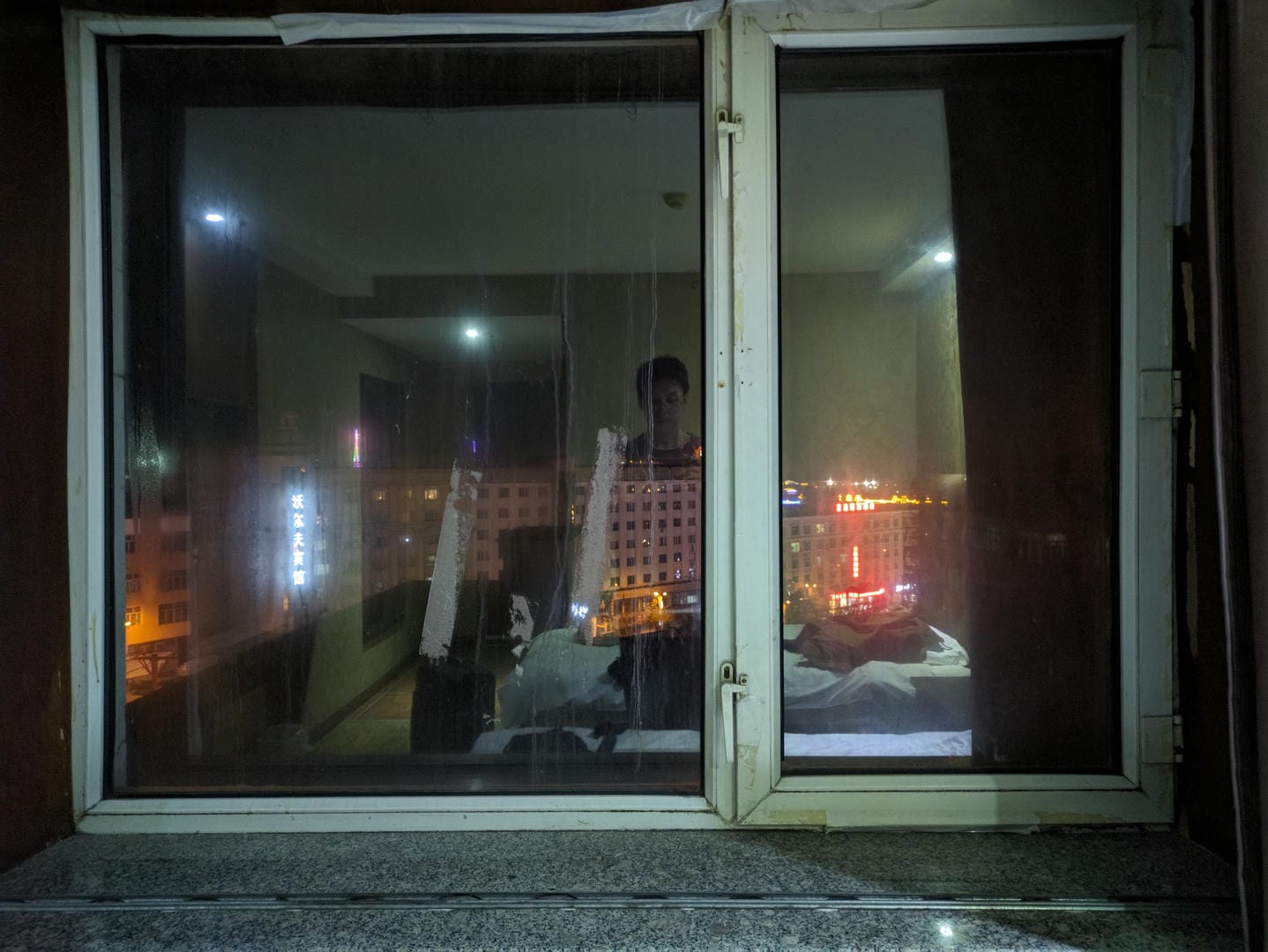 Through a window, a person is seen lying on a bed inside a room, with the city’s night view outside.