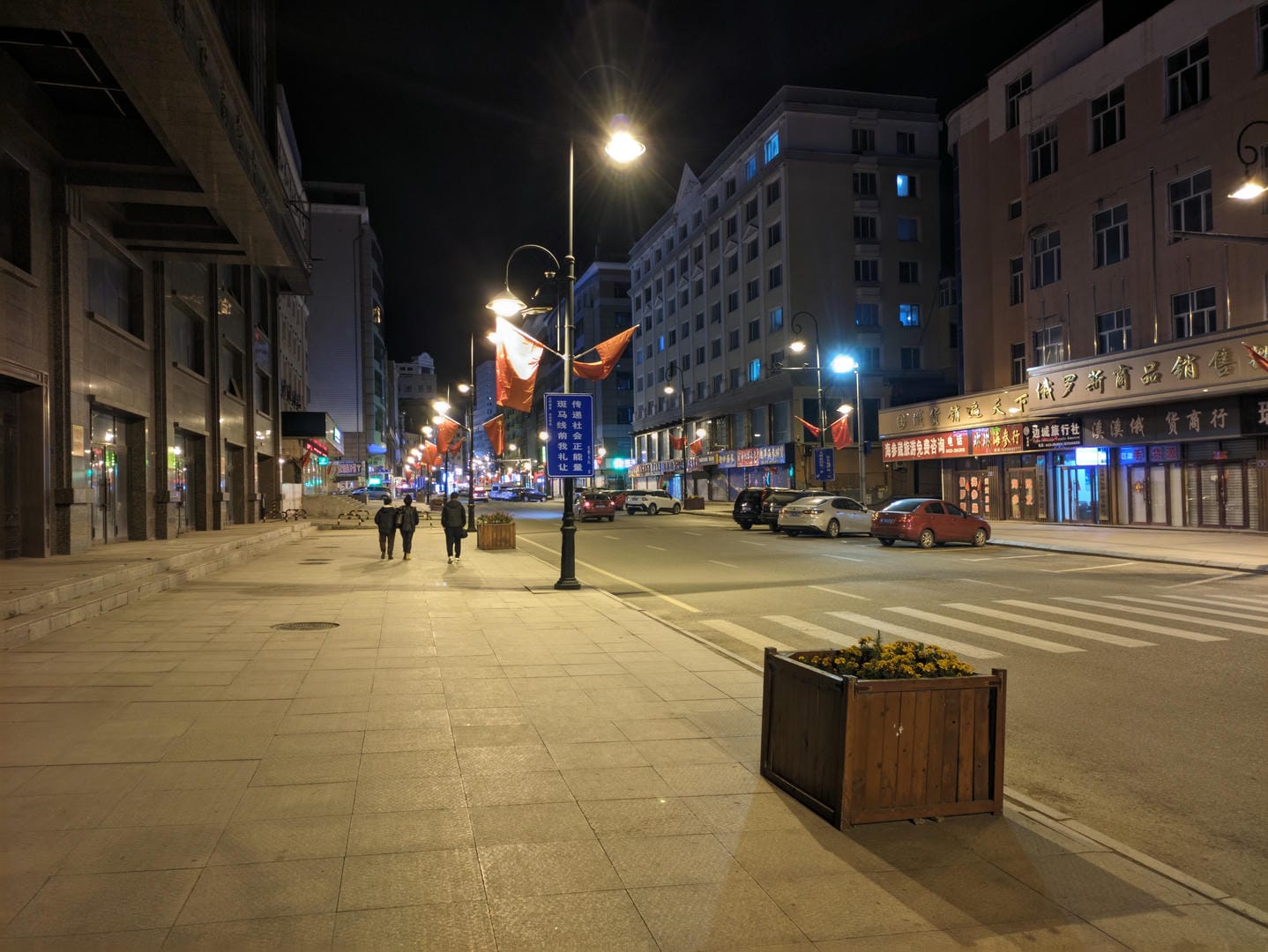 A city street at night, streetlights on, with two people walking on the sidewalk.