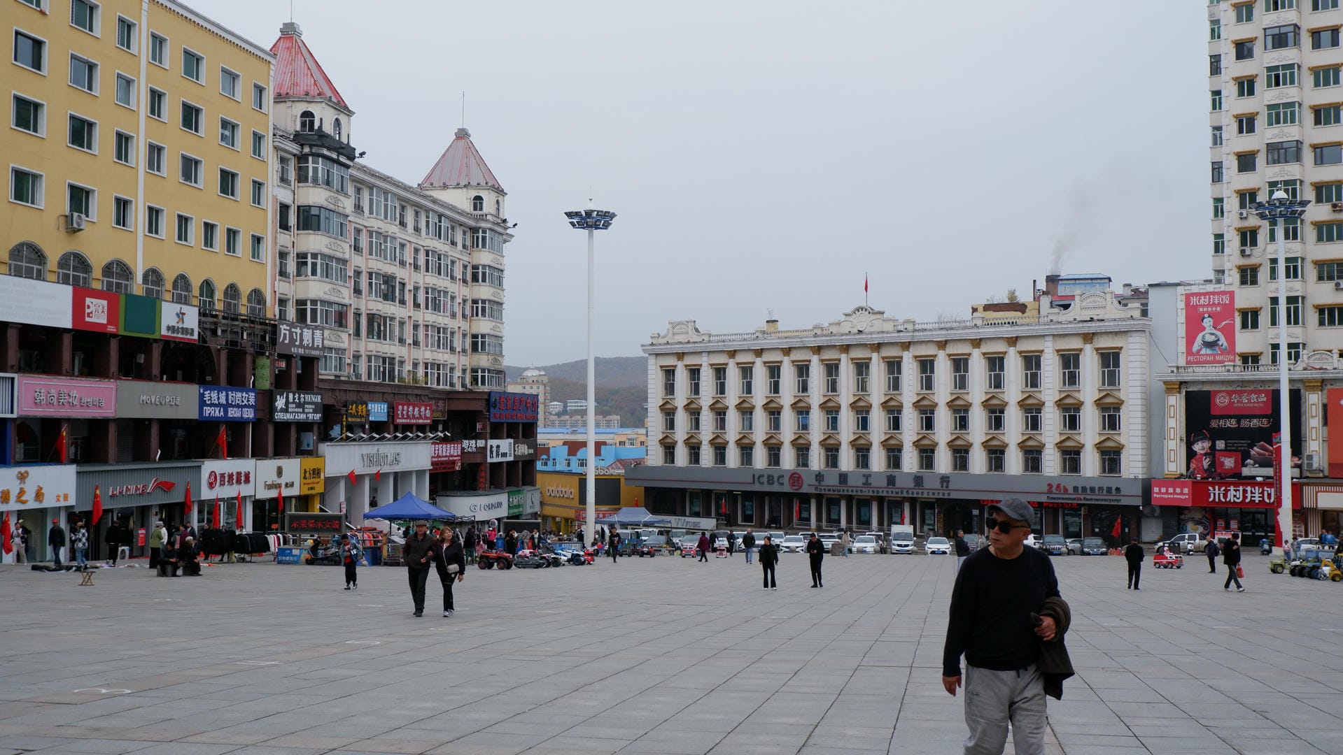 A square surrounded by several buildings, including a white and yellow building. There are a few people walking around in the square.