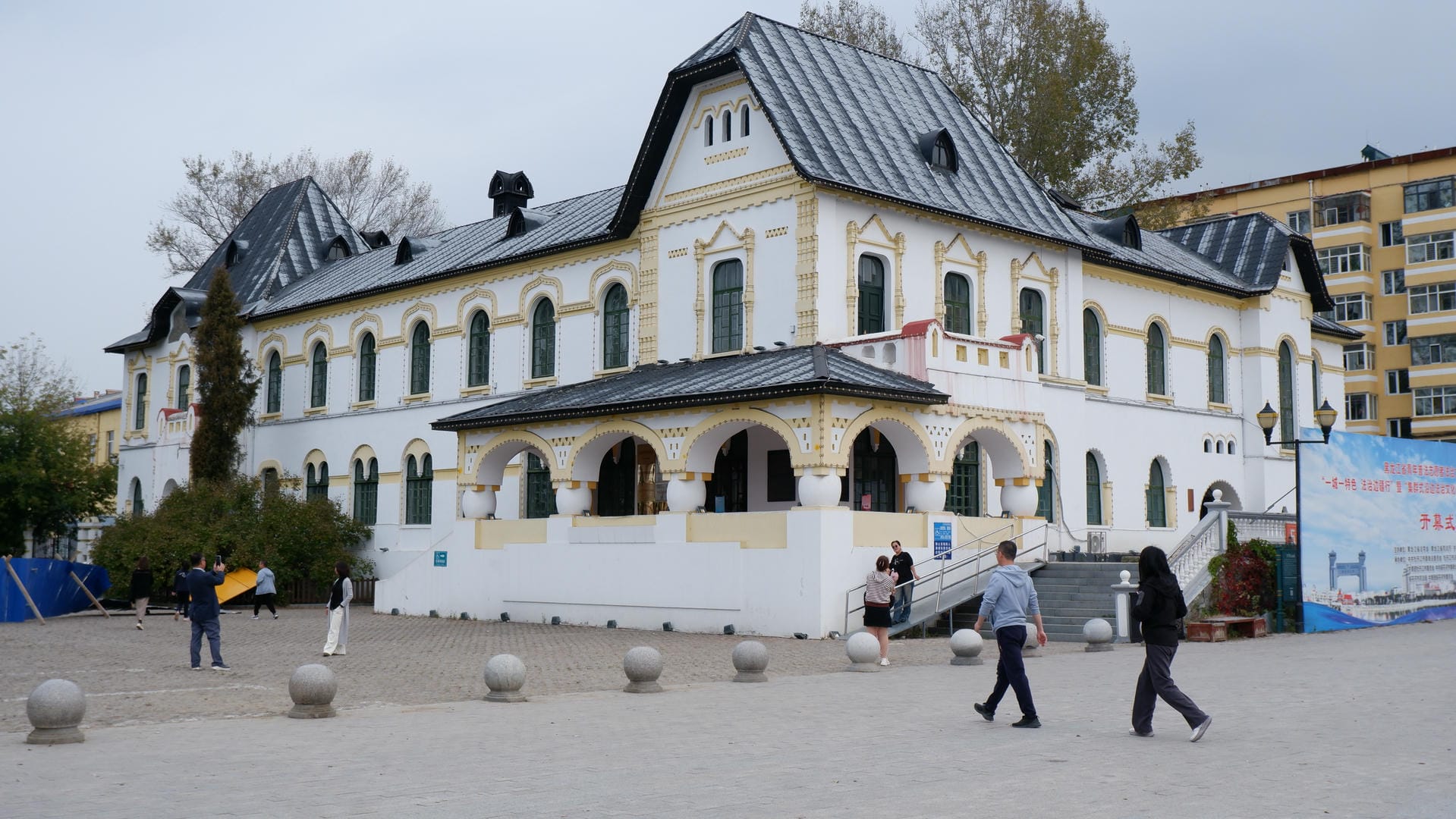 A white building with a black roof. In front of the building, there is a small square where some people are active.
