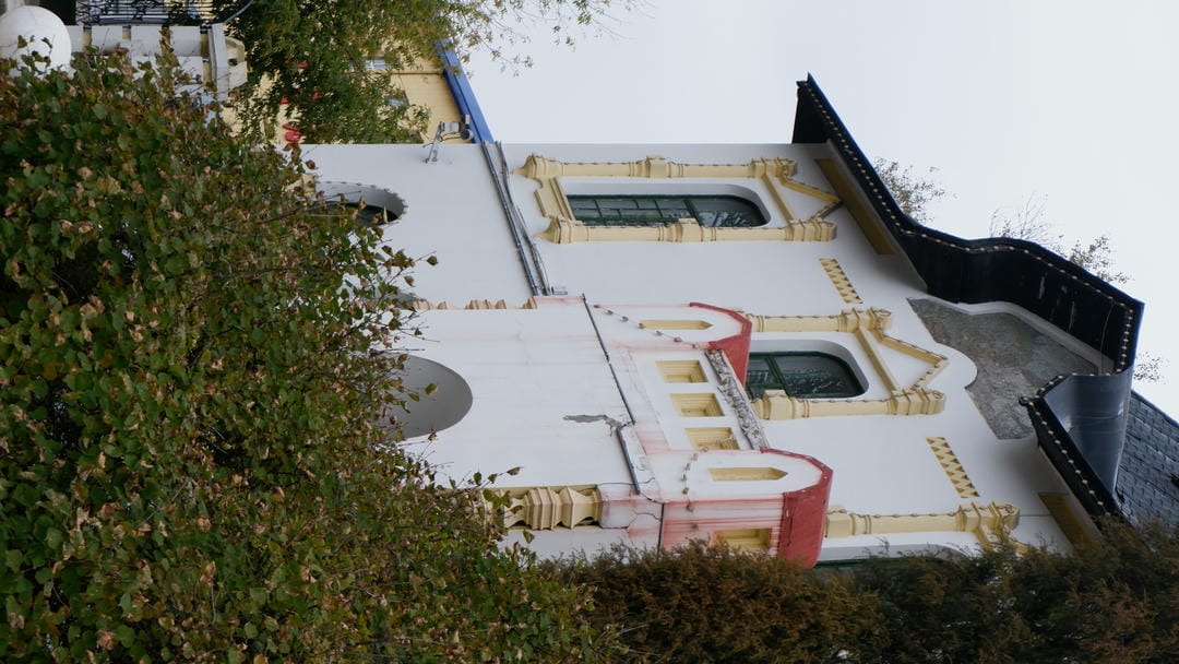 A section of a white building with unique architectural style, featuring beautifully decorated windows and some green plants in the foreground.
