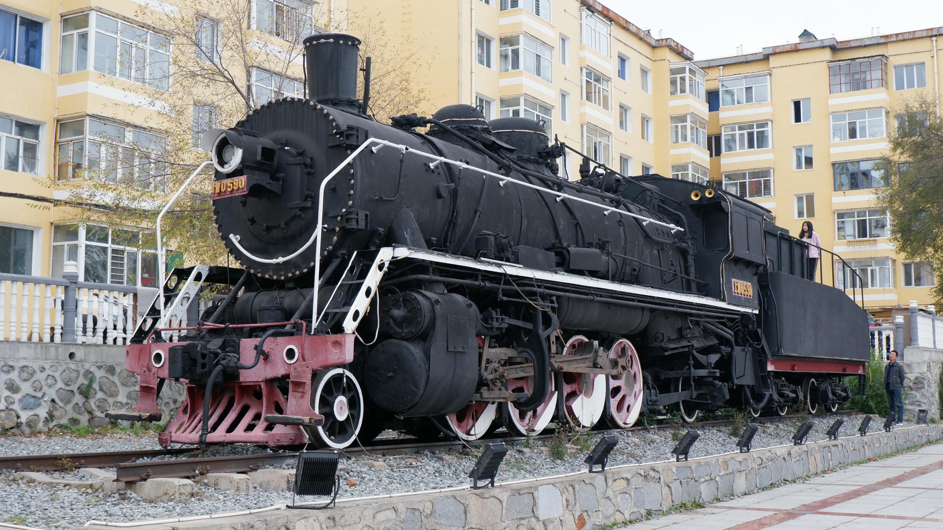 A black steam locomotive parked on the tracks, with red decorations in front and yellow buildings in the background.