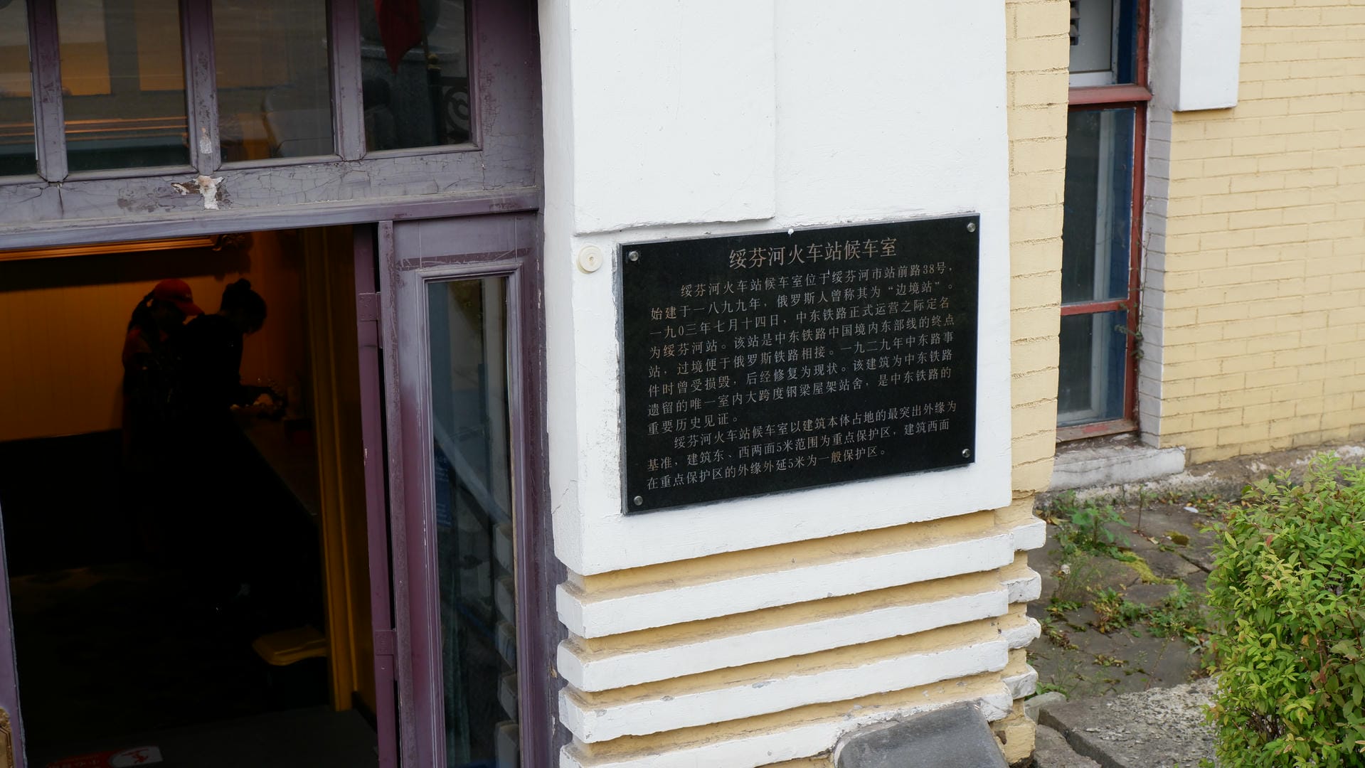 Next to a door, there is a black metal plaque inscribed with historical information about the Suifenhe Railway Station waiting room.