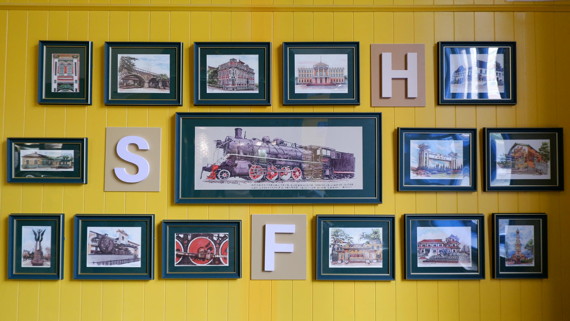 Multiple framed photos hang on a yellow wall. In the center is a painting of a steam train, surrounded by photos of buildings, with the letters “SHF” formed on the wall.