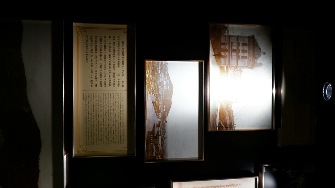 Two old photographs hang on the wall, with a rectangular explanatory plaque below them, illuminated by light from above.