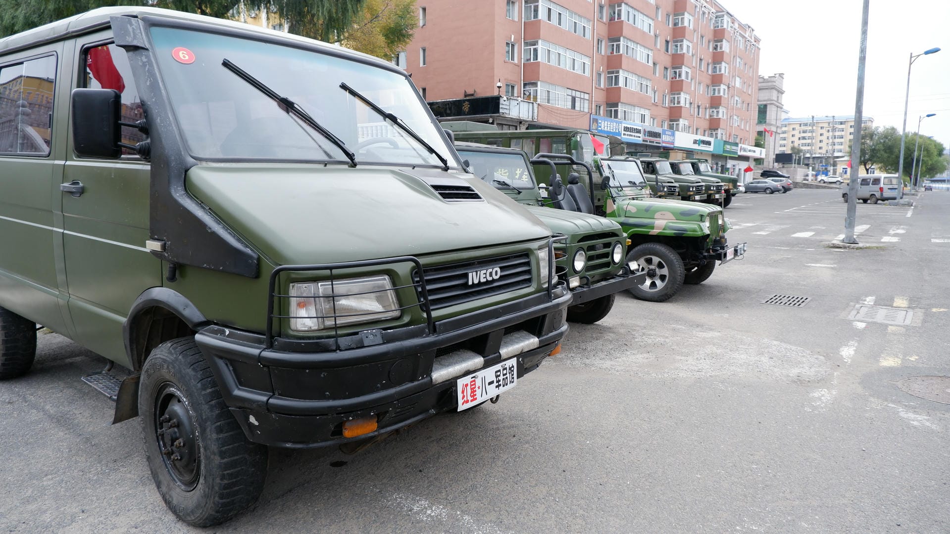 A row of green military vehicles, including vans and trucks, are parked in a lot.