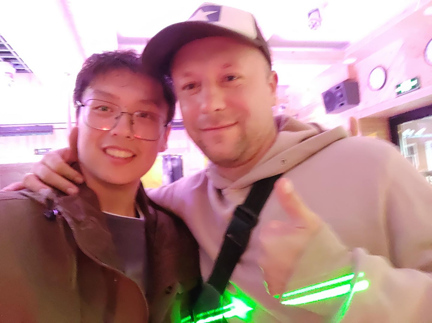 Two people take a selfie indoors, with pink lights and decorations in the background.