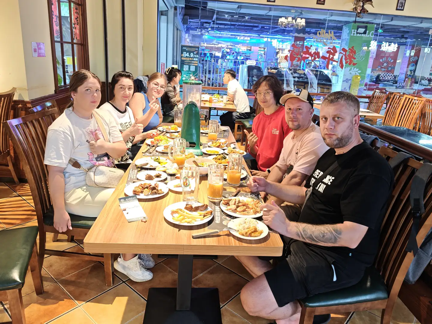 A group of people are sitting around a long table in a restaurant, with food and drinks laid out on the table, including french fries, meat dishes, and orange juice. They are dressed in casual attire, some smiling and others gesturing; in the background, you can see other customers and the shop's signboard. The interior of the restaurant is warmly decorated, featuring wooden tables and chairs as well as glass windows, through which you can see a bright commercial street scene.