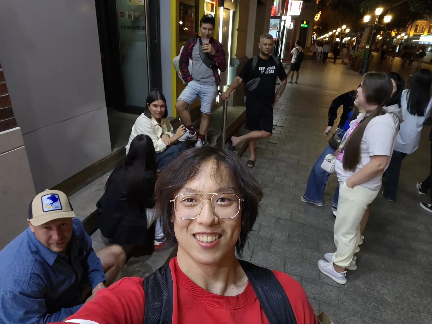 A man wearing glasses is taking a selfie on the street. He is wearing a red top and carrying a black backpack, while several people are standing or sitting around him. The background is a nighttime city street, with lights illuminating pedestrians and shops, and the ground paved with stone bricks. People are dressed in summer clothes; some are chatting, and others are looking at their phones.