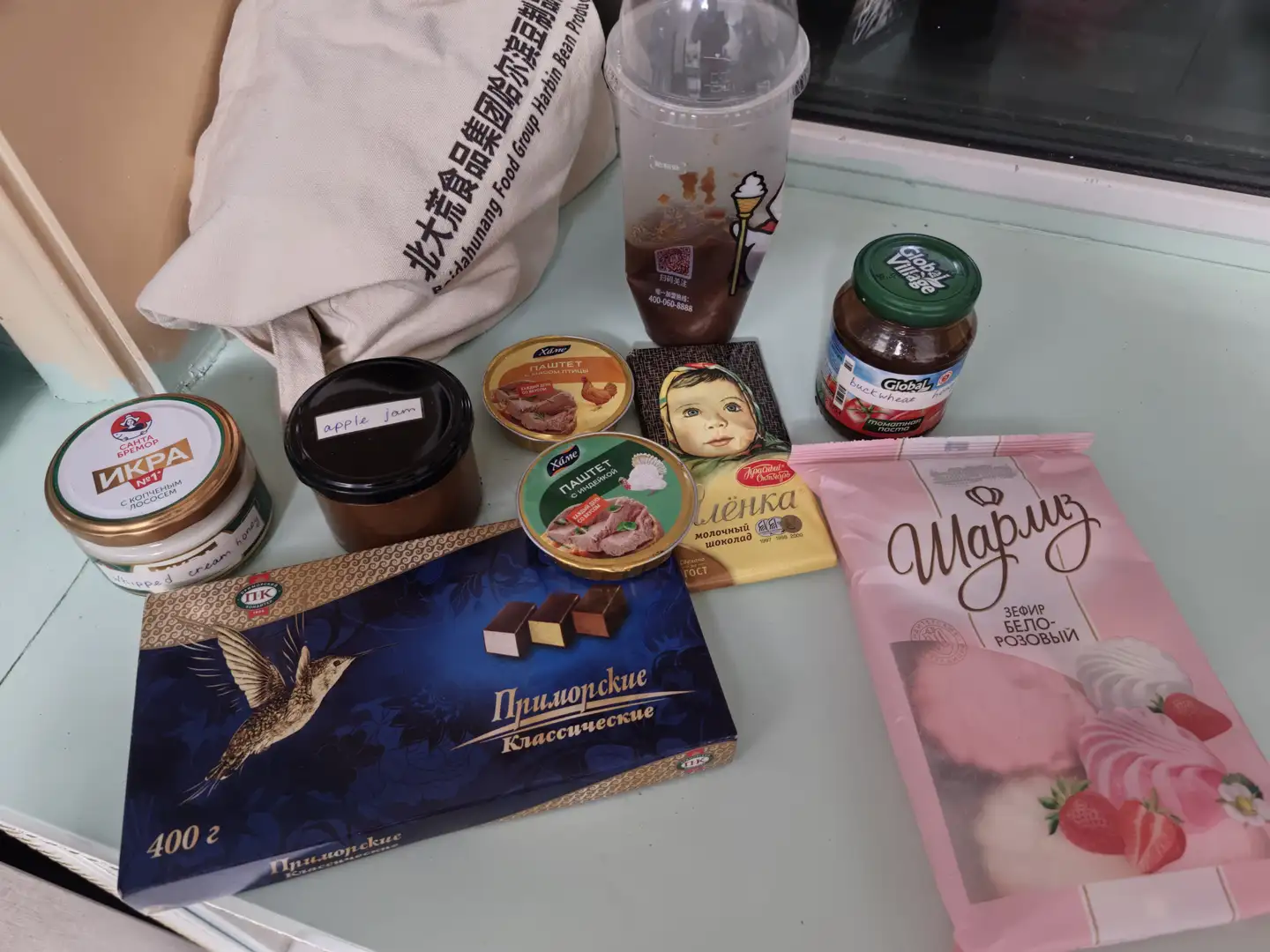 On the desktop are various food items, including a box of chocolates printed with flying birds, a bag of pink-packaged candies, several jars of jam and meat products, a cup of milk tea, and a cloth bag printed with Chinese characters. The chocolate packaging has the Russian text “Приморские Классические” while the candy packaging reads “Марица” and “Зефир Бело-Розовый”. Nearby are jars labeled “apple jam” and a jam bottle marked “Global Village”.